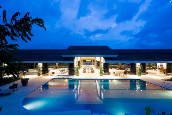 A luxurious resort-style building with an illuminated swimming pool is pictured under a blue, evening sky.