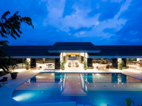 A luxurious resort-style building with an illuminated swimming pool is pictured under a blue, evening sky.