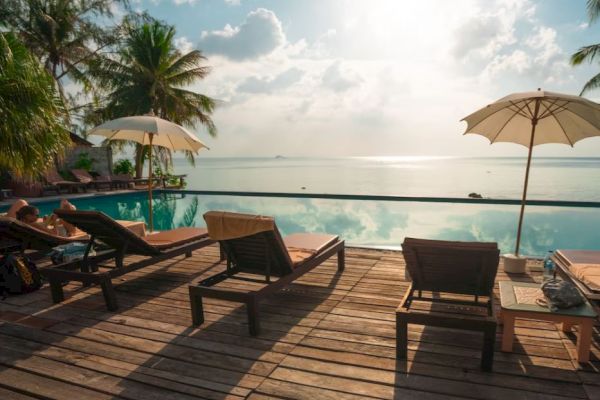 Lounge chairs and umbrellas by a serene pool with palm trees and an ocean view under a partly cloudy sky.