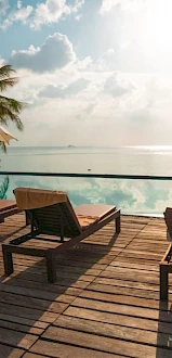 A serene poolside scene with deck chairs, umbrellas, palm trees, and ocean view under a partly cloudy sky at sunset.