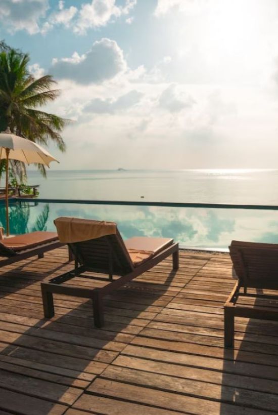 A serene beachfront scene with lounge chairs, parasols, and a tranquil pool, overlooking the ocean under a partly cloudy sky.