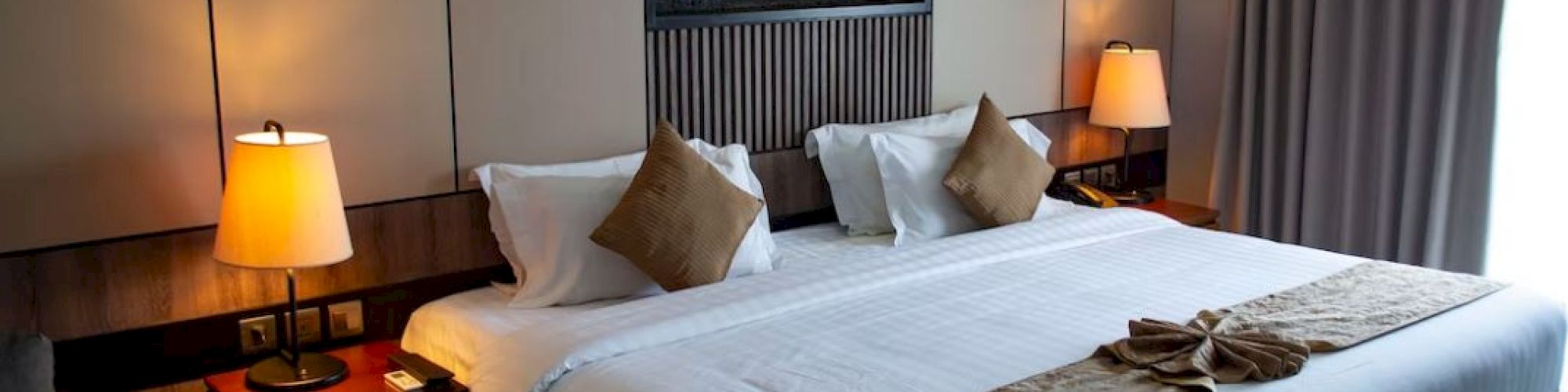 A cozy hotel room features a large bed with pillows, bedside lamps, and curtains, creating a warm and inviting atmosphere.