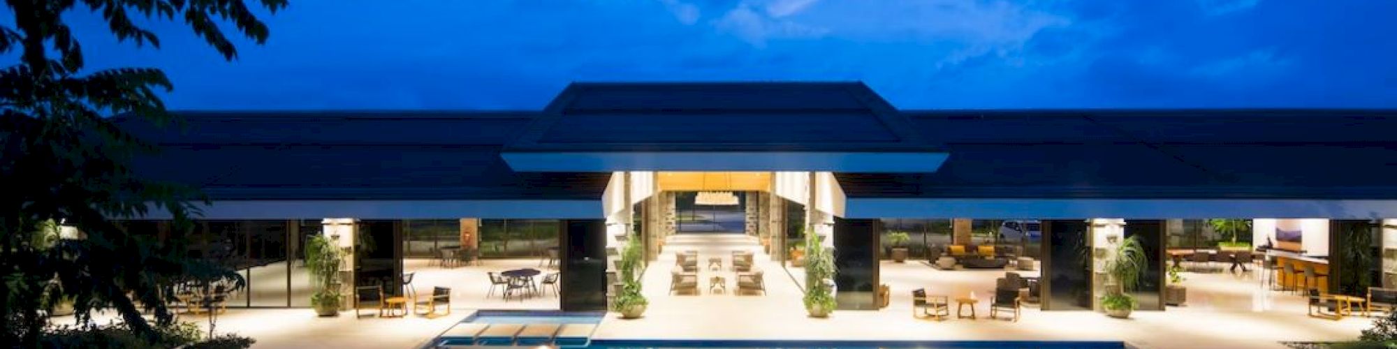 The image shows a luxurious modern building with a large illuminated swimming pool under a twilight sky.