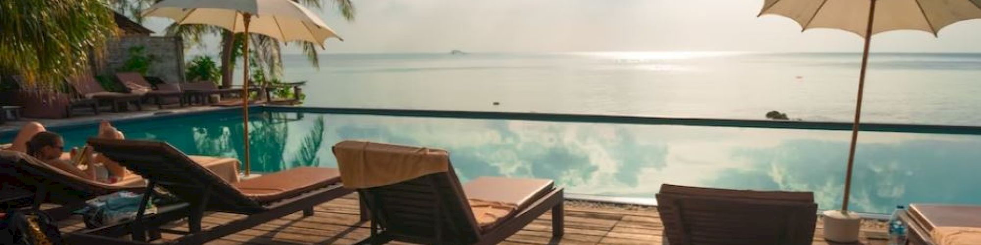 A serene poolside scene with lounge chairs, palm trees, and sun umbrellas overlooking a calm body of water under a cloudy sky.