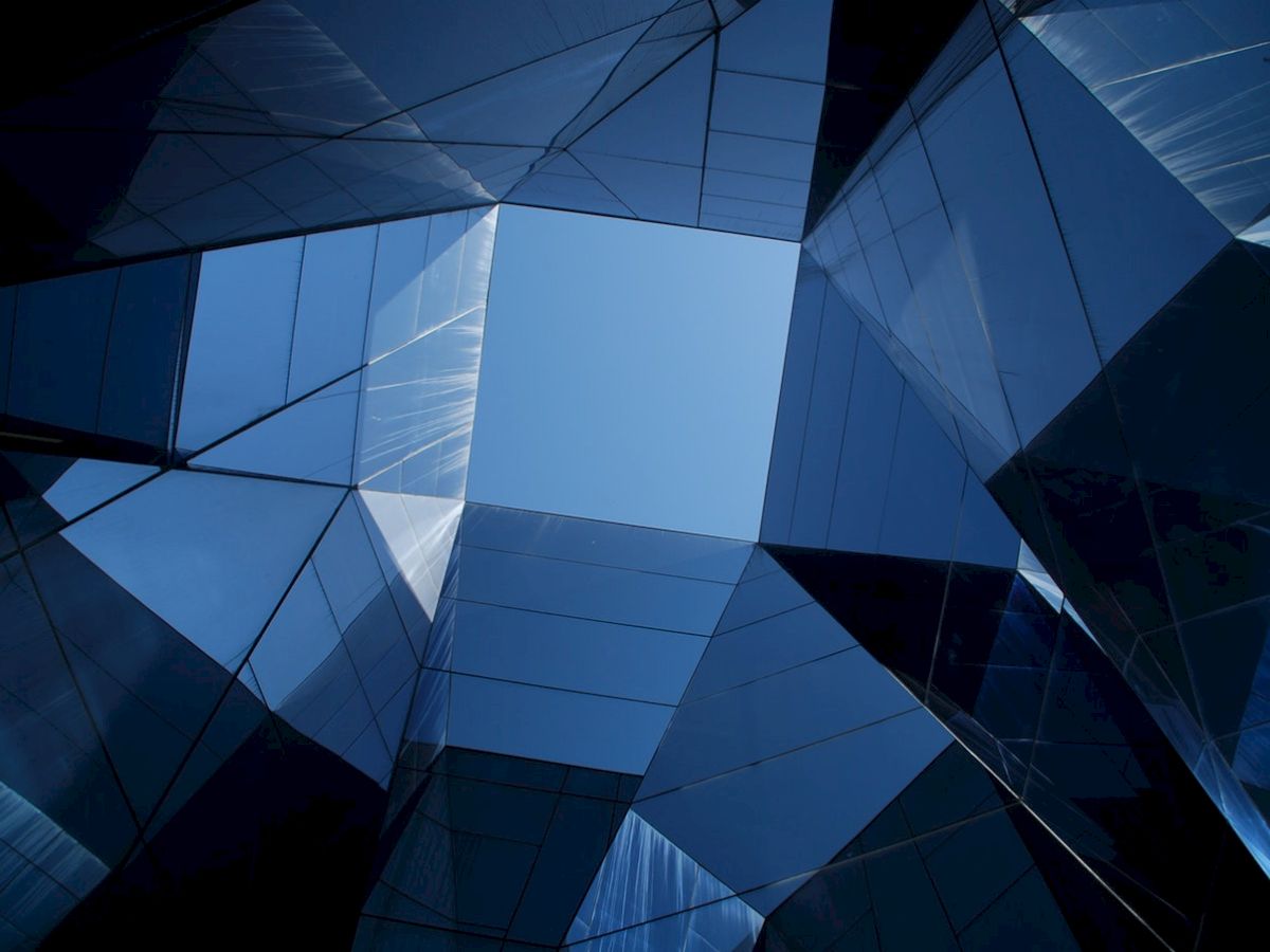 The image shows a view looking up at angular, reflective modern architecture with a clear blue sky visible through the geometric shapes.