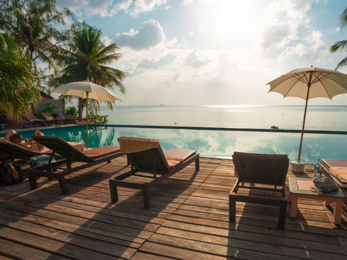 A serene poolside scene with sunbeds, parasols, palm trees, and a view of a calm ocean under a partly cloudy sky.