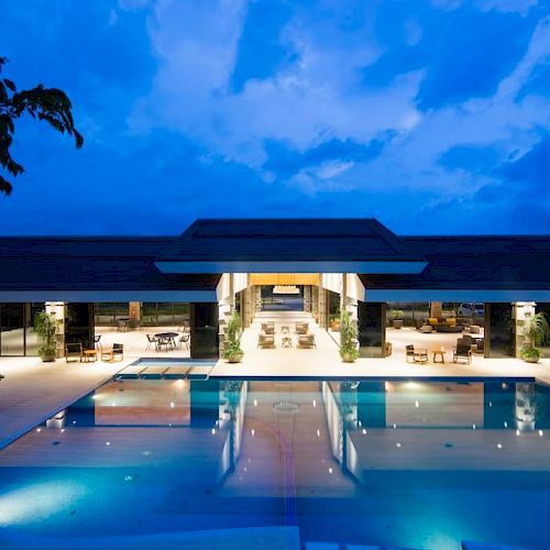 A luxurious building with a large, illuminated pool in the foreground, surrounded by seating areas and set against a vibrant evening sky.