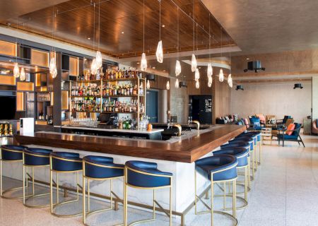 A modern bar with a wooden counter, blue barstools, hanging lights, and a well-stocked array of drinks. Cozy seating is visible nearby.