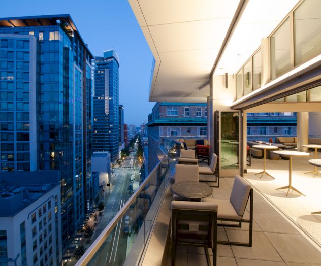 A modern balcony with seating overlooks a city street and tall buildings at dusk, featuring a spacious indoor-outdoor design.