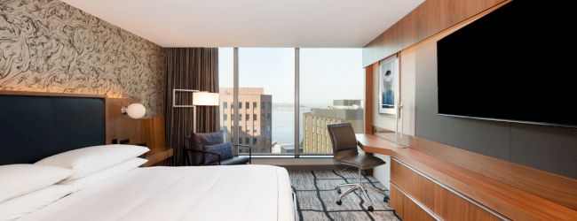 A modern hotel room with a large bed, wall-mounted TV, desk, chair, and a window view of buildings and water in the background.