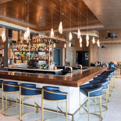 A modern bar with blue stools, pendant lights, and a well-stocked shelf of bottles. Casual seating is in the background.