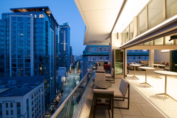 A modern rooftop terrace with seating overlooks a cityscape featuring tall glass buildings, viewed during twilight hours.