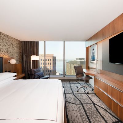 A modern hotel room with a large bed, TV, desk, and window view of buildings and water.