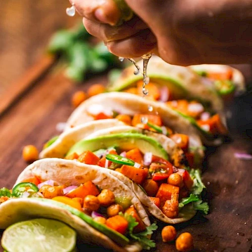 A hand is squeezing lime over tacos filled with chickpeas, avocado, onions, and cilantro. Lime slices are placed in front on a wooden surface.