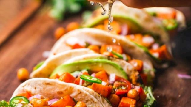 A hand is squeezing lime over tacos filled with chickpeas, avocado, onions, and cilantro. Lime slices are placed in front on a wooden surface.