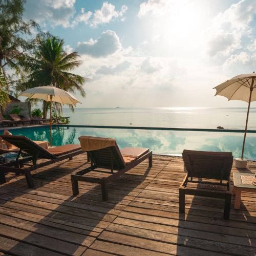 Beachfront scene with lounge chairs, umbrellas, a pool, and palm trees under a bright, partly cloudy sky reflecting in the water.