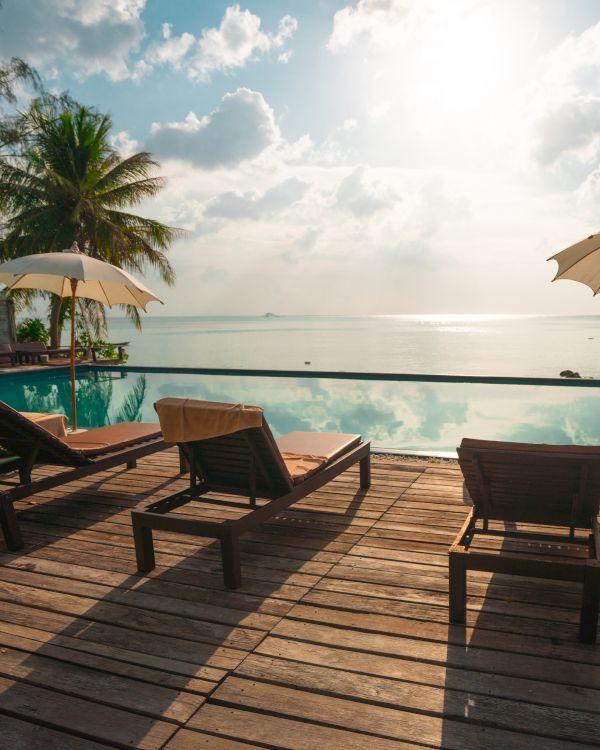 Beachfront scene with lounge chairs, umbrellas, a pool, and palm trees under a bright, partly cloudy sky reflecting in the water.