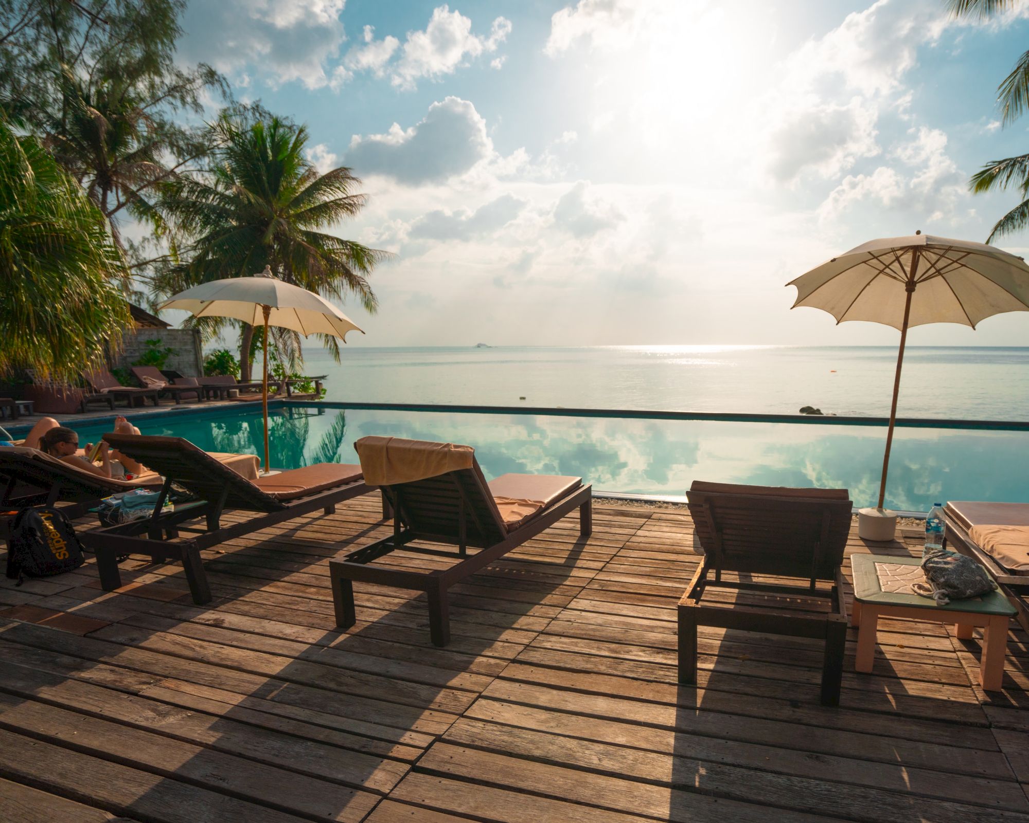 Lounge chairs and umbrellas by a pool overlook a calm sea and sunset, with a wooden deck and tropical foliage surrounding the area.