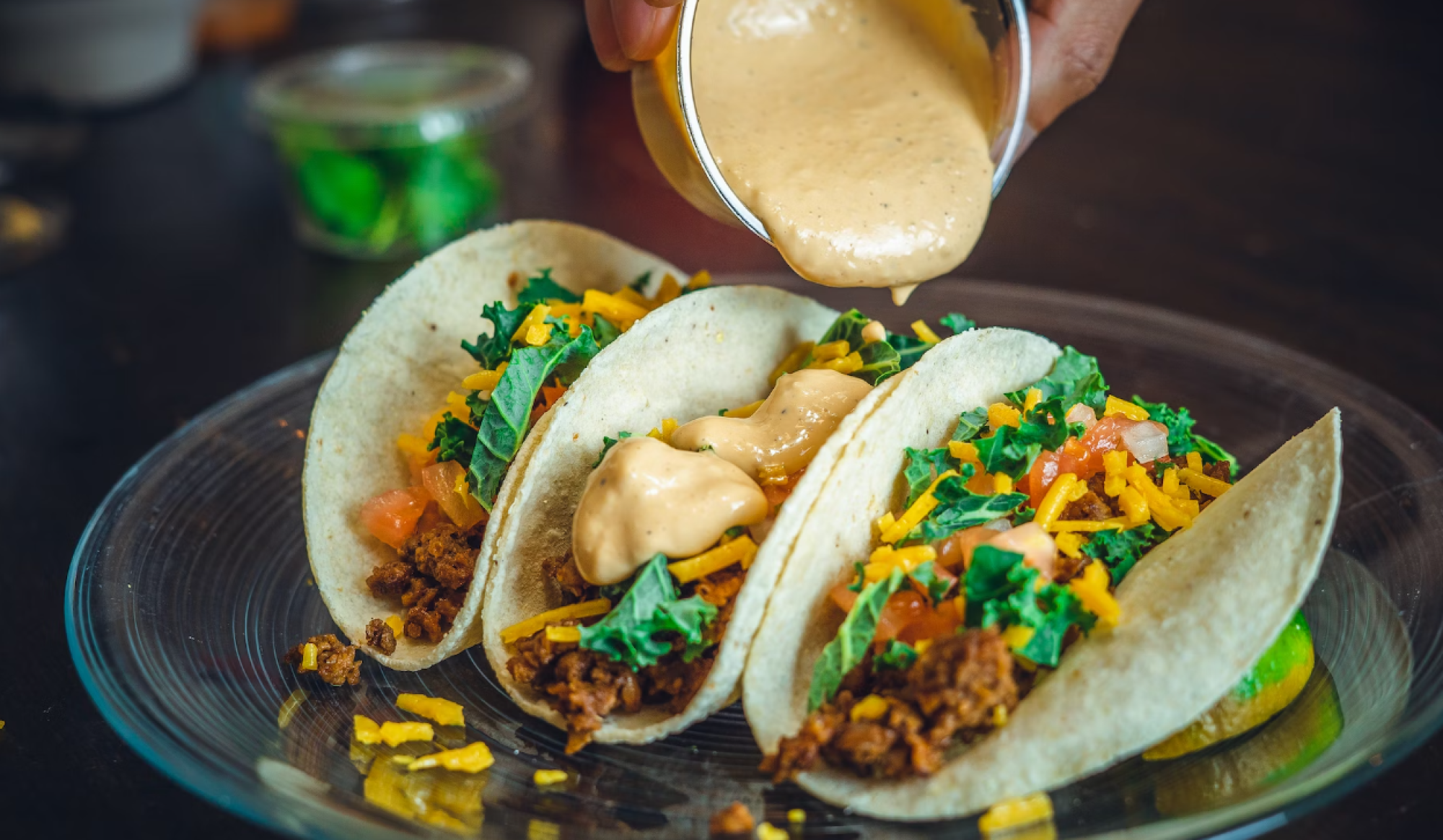 Three tacos with ground meat, cheese, lettuce, and sauce being poured over them on a transparent plate.
