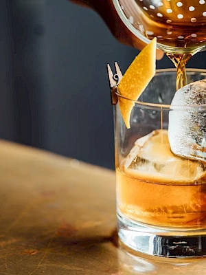 A hand is pouring a drink into a glass with ice and a citrus twist, placed on a wooden surface.