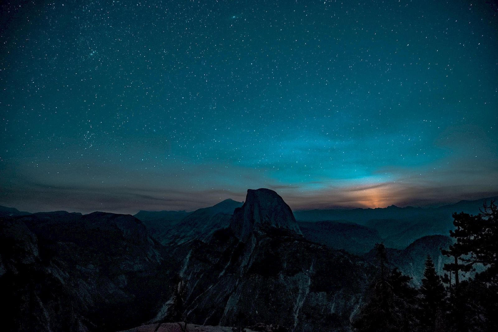A starry night sky over a mountainous landscape with a prominent peak and a glowing horizon, creating a serene, dark silhouette.