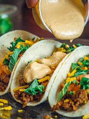 Three tacos with meat, cheese, and greens, topped with a creamy sauce being poured from a cup.