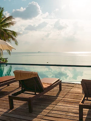 A serene poolside scene with lounge chairs, umbrellas, and a stunning ocean view, bordered by palm trees under a partly cloudy sky.