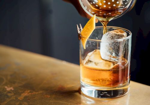A hand pours a brown cocktail from a strainer into a glass with ice, garnished with a twist of orange peel.