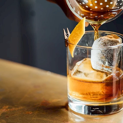 A hand pours a brown cocktail from a strainer into a glass with ice, garnished with a twist of orange peel.
