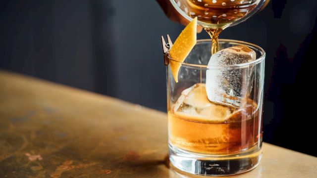 A hand pours a brown cocktail from a strainer into a glass with ice, garnished with a twist of orange peel.