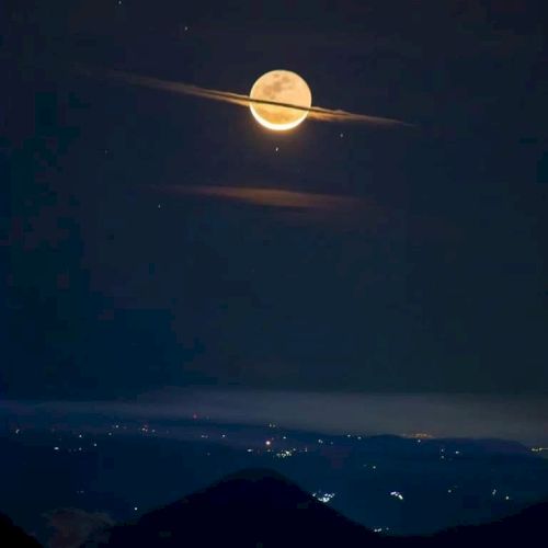 A bright full moon appears with a ring of clouds, resembling Saturn, over a landscape with distant city lights in the darkness.