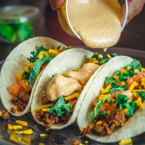 Three tacos with meat, cheese, lettuce, and tomatoes on a plate, being topped with a creamy sauce.