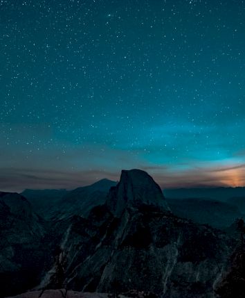 A starry night sky over a mountainous landscape, with a glowing horizon and silhouettes of trees in the foreground.