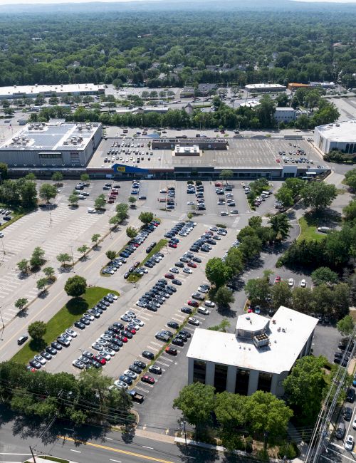Aerial view of a large parking lot with numerous cars, surrounded by greenery, roads, and various buildings in the background.