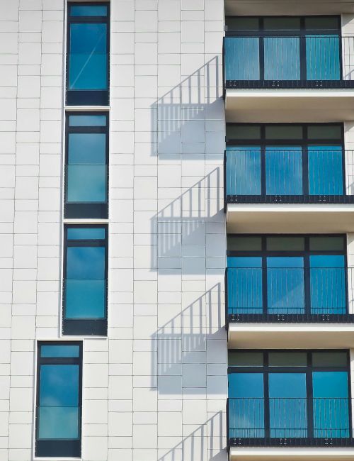 A modern building facade with white tiles, tall blue-tinted windows, and balconies, set against a clear blue sky with some clouds.