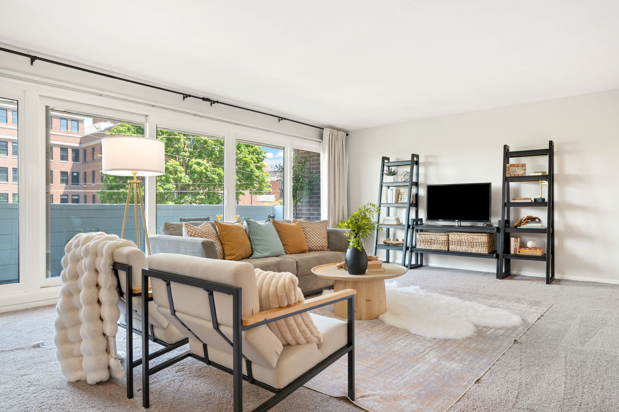 A bright living room with modern furniture, a large window, shelves, a TV, and cozy decor elements like cushions and a throw blanket.