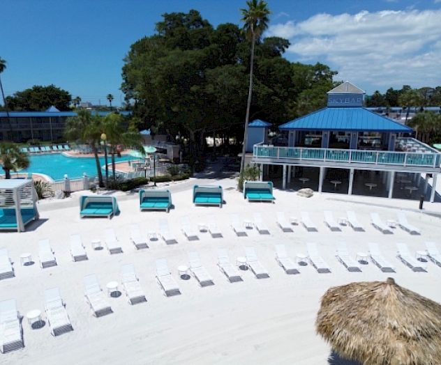 A beach resort with white sand, lounge chairs, cabanas, a pool, and a nearby bar under a clear blue sky offers a tranquil setting.