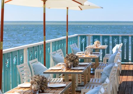 A scenic outdoor dining area by the sea with wooden tables, white chairs, umbrellas, and decorative floral centerpieces on a sunny day.