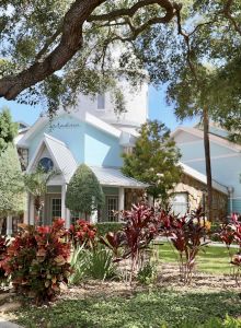 A building with a blue exterior surrounded by lush greenery, trees, and a pathway under sunlight.