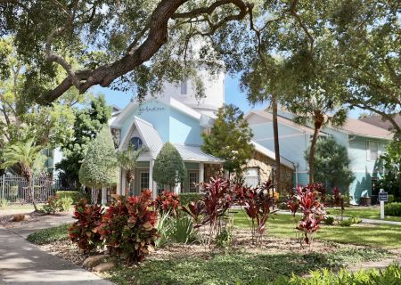 A building with a blue exterior surrounded by lush greenery, trees, and a pathway under sunlight.