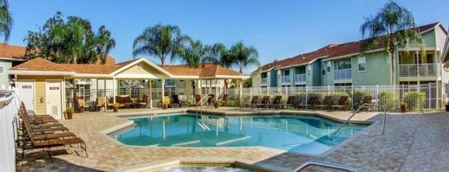 A serene outdoor pool area with lounge chairs, a hot tub, surrounded by buildings and palm trees under a clear blue sky.