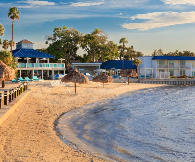 A sandy beach with palm trees, a resort, thatched umbrellas, and calm waters under a clear sky.