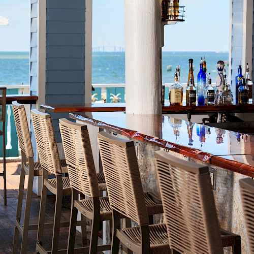 Outdoor bar with wooden chairs, overlooking a body of water. Umbrella-shaded tables and blue skies create a relaxed atmosphere.