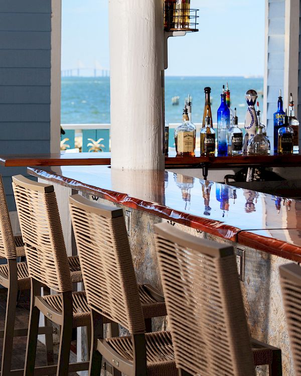 Outdoor bar with wooden chairs, overlooking a body of water. Umbrella-shaded tables and blue skies create a relaxed atmosphere.