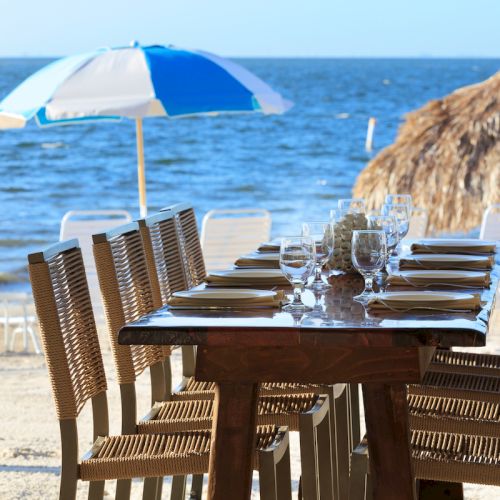 A beachside dining setup with a table, chairs, and place settings, under umbrellas near the ocean, with thatched huts in the background.