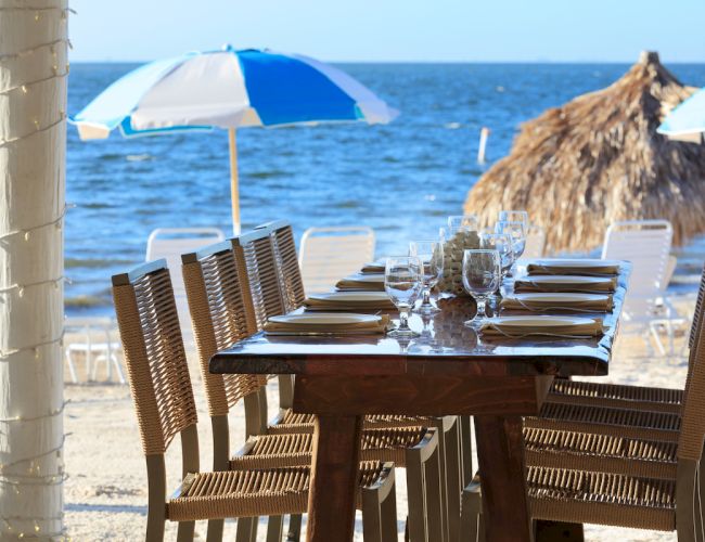 A beachside dining setup with a table, chairs, and place settings, under umbrellas near the ocean, with thatched huts in the background.