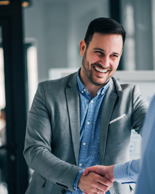 Two men in business attire are shaking hands in an office setting, appearing to be in a friendly and professional interaction.