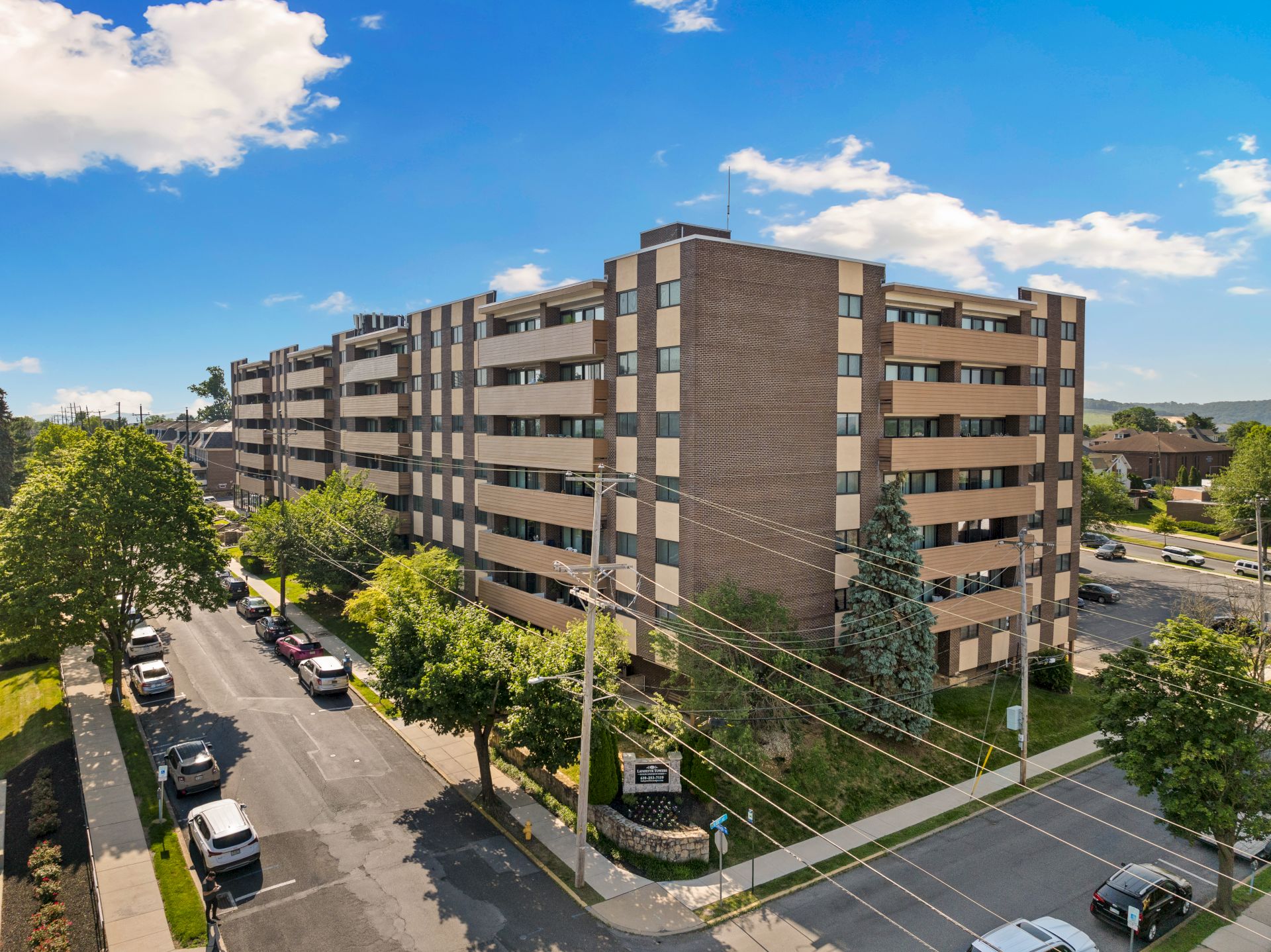 The image shows a multi-story residential building with parked cars, trees, and a clear blue sky above, creating a suburban scene.