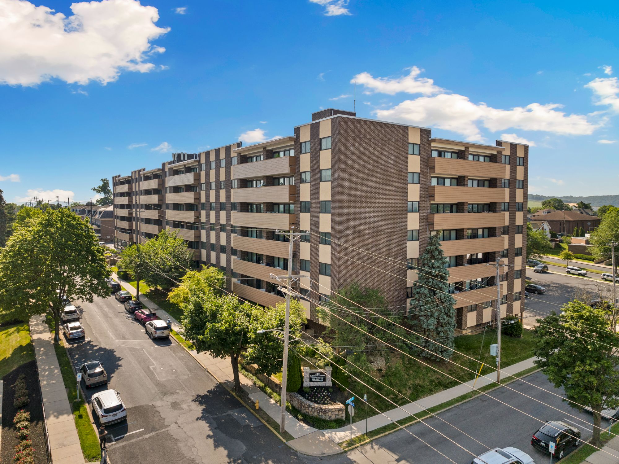 The image shows a multi-story residential building with parked cars, trees, and a clear blue sky above, creating a suburban scene.