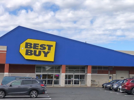 The image shows a Best Buy store with a blue facade, large yellow sign, and a parking lot with several cars.